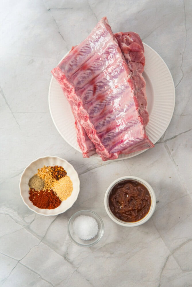 Raw pork ribs on a white plate, surrounded by small bowls containing mixed spices, coarse salt, and barbecue sauce, set on a light marble surface.
