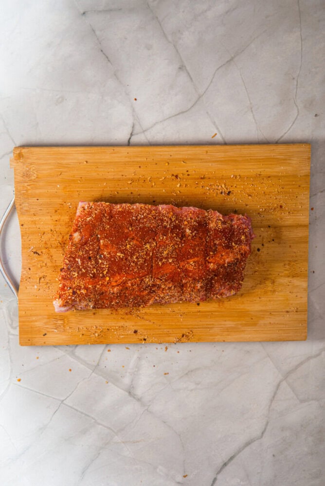 Raw rack of ribs coated with seasoning resting on a wooden cutting board, placed on a light-colored marble surface.