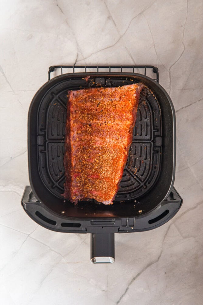 A seasoned rack of ribs sits inside an air fryer basket on a light-colored marble surface.