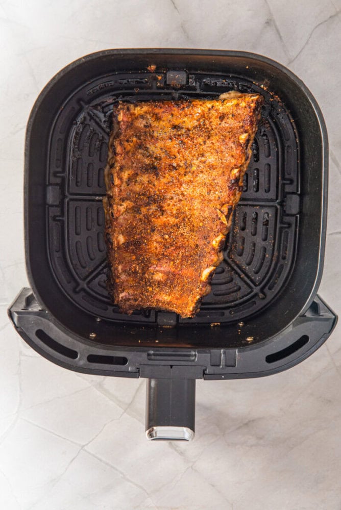 A rack of seasoned ribs cooked in an air fryer basket sits on a light-colored countertop.