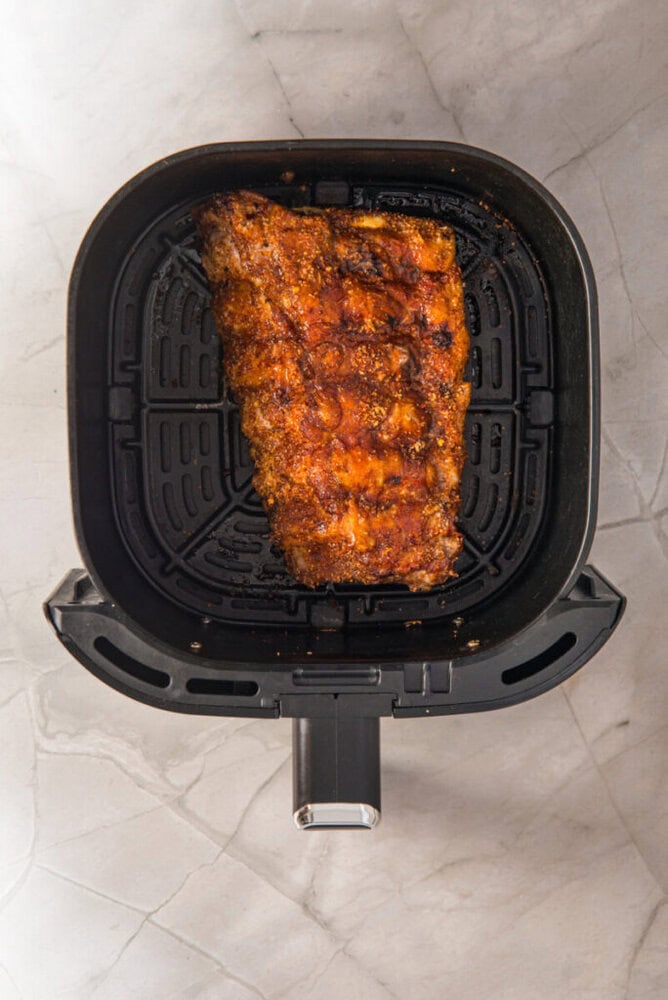 A rack of seasoned, cooked ribs sits inside an air fryer basket on a marble countertop.
