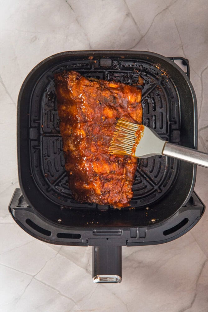 A rack of glazed ribs is being brushed with sauce inside an air fryer basket on a light-colored countertop.