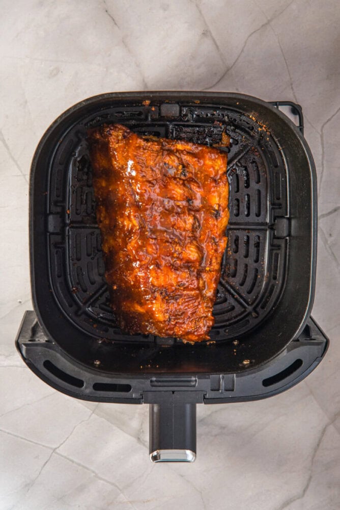 A slab of glazed ribs sits inside the basket of an air fryer on a gray marble surface.