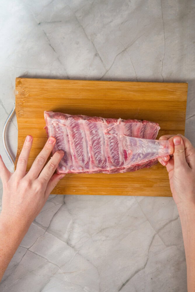 A person removes the membrane from a rack of raw pork ribs on a wooden cutting board placed on a marble countertop.