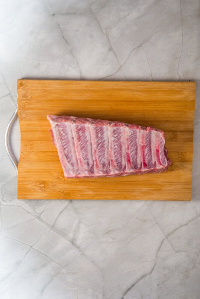 A raw rack of pork ribs on a wooden cutting board atop a light gray marble surface.