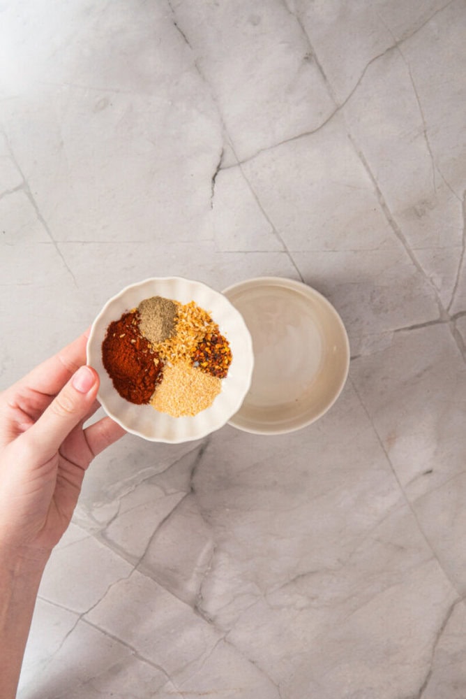 A hand holds a small white bowl containing assorted ground spices over an empty bowl on a light marble surface.