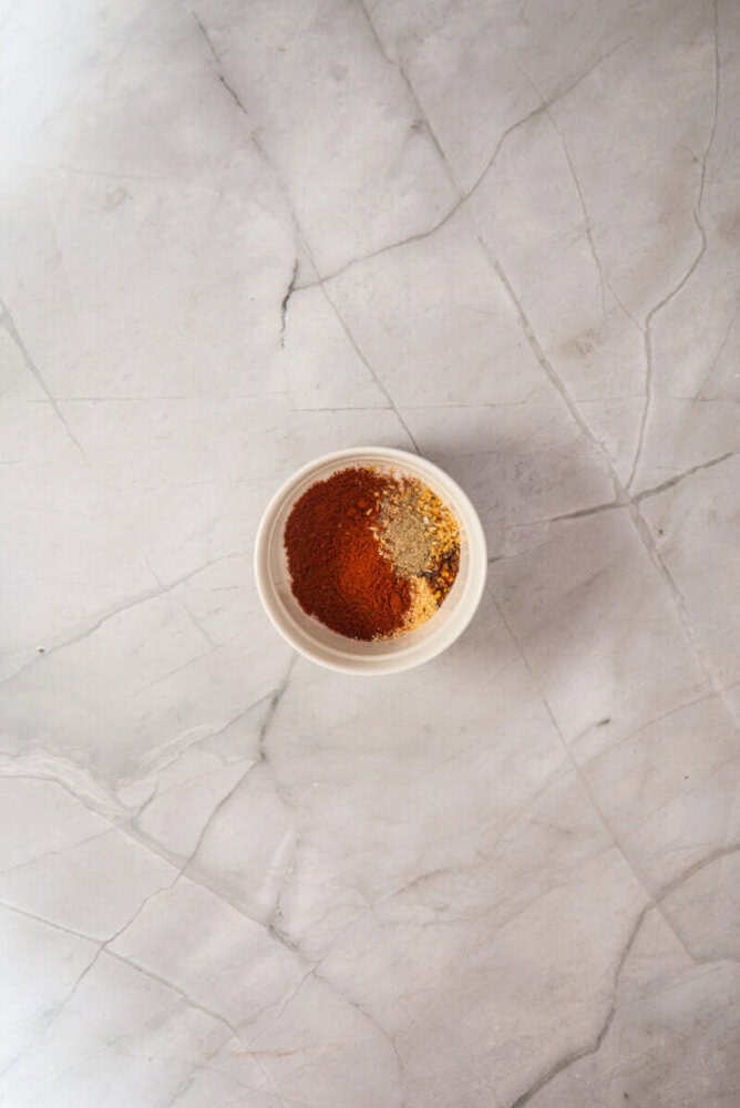 A small white bowl containing a mix of ground spices sits on a light gray marble surface.