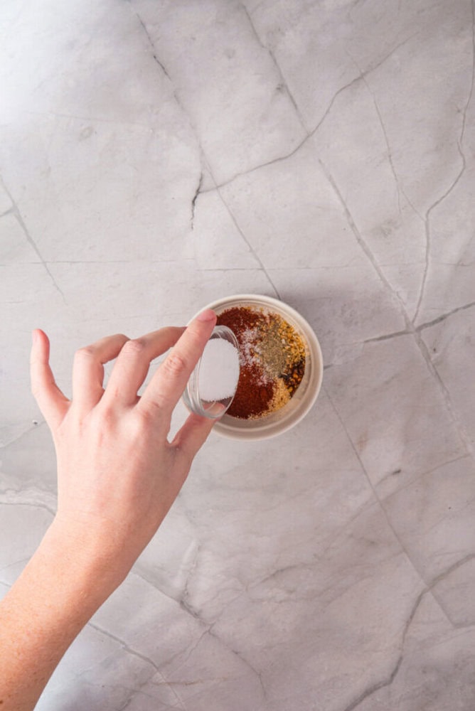 A hand pours salt from a small glass bowl into a larger bowl containing mixed spices on a marble countertop.