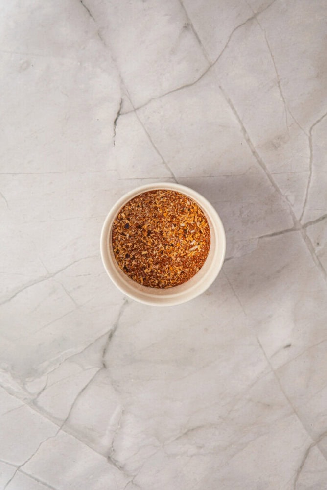 A small white bowl filled with a reddish-brown spice blend sits on a light gray, marbled surface.