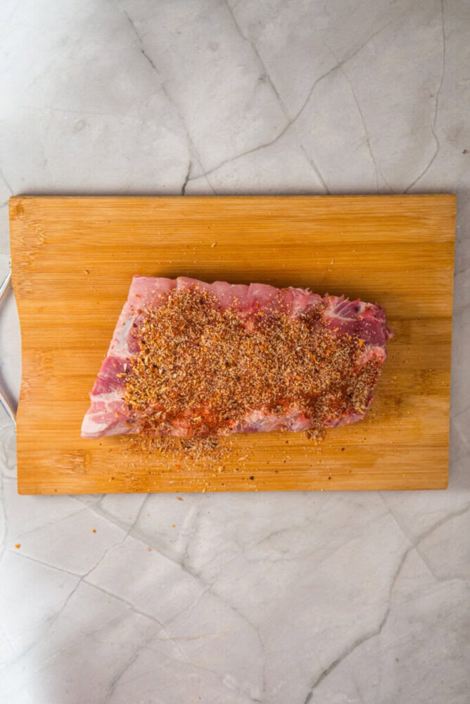 A slab of raw meat coated with dry seasoning sits on a wooden cutting board on a marble countertop.