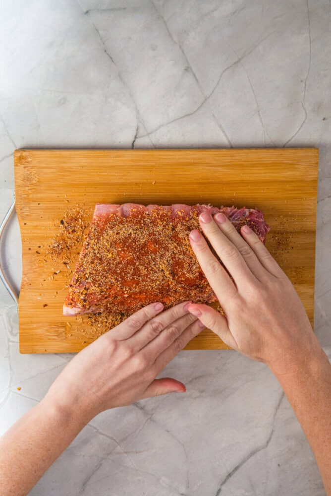 Two hands rub seasoning onto a rack of raw ribs placed on a wooden cutting board over a marble surface.