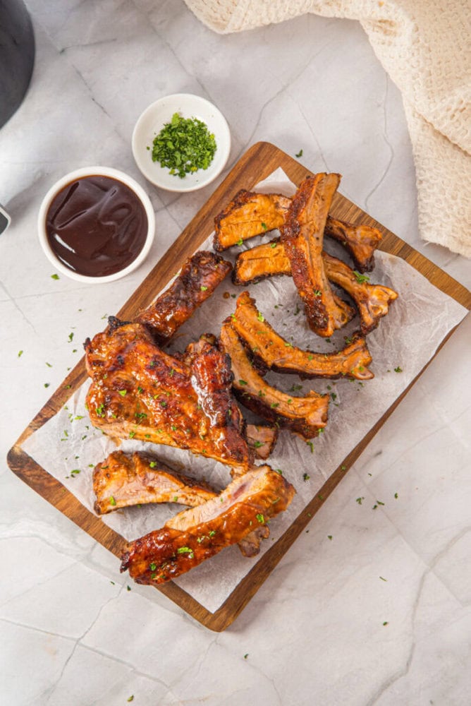 A wooden board with cooked pork ribs garnished with herbs, served alongside a small bowl of barbecue sauce and a dish of chopped herbs on a marble surface.
