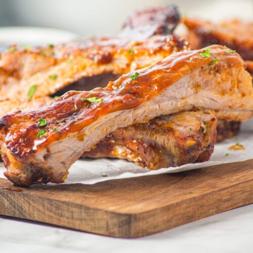 Barbecue pork ribs stacked on a wooden cutting board, garnished with chopped herbs, with a small bowl of sauce in the background.