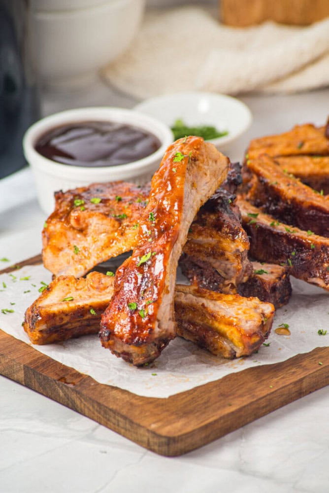A stack of cooked pork ribs garnished with herbs on a wooden board, with a bowl of barbecue sauce in the background.