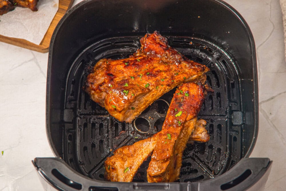 Two cooked pork ribs with seasoning rest in the basket of an air fryer on a light-colored countertop.