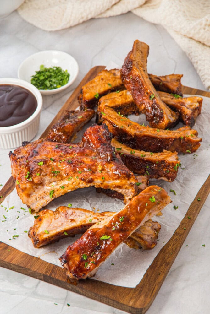 A wooden board with glazed pork ribs garnished with herbs, accompanied by a small bowl of barbecue sauce and chopped chives on the side.