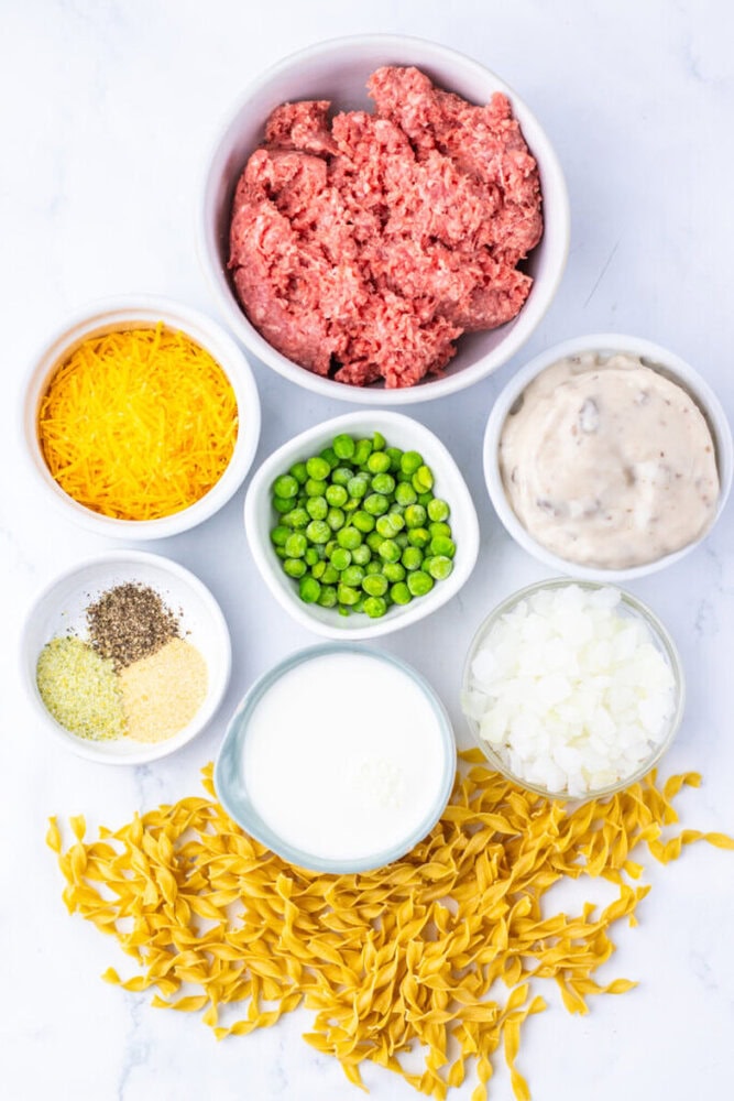 Bowls containing ground beef, shredded cheddar cheese, peas, cream soup, chopped onions, milk, seasonings, and uncooked egg noodles arranged on a white surface.