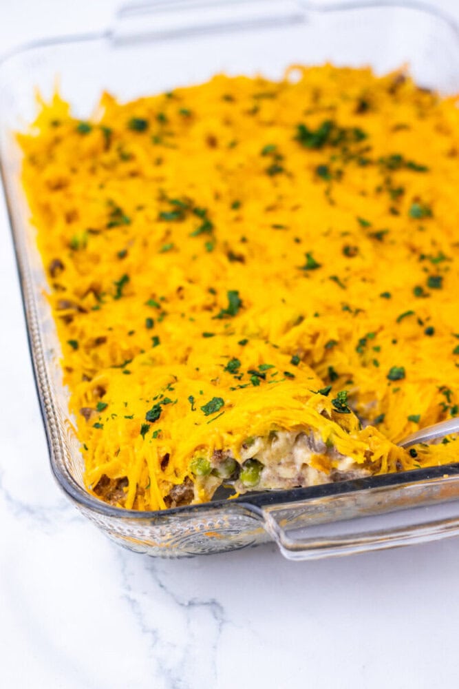 A glass baking dish with a casserole topped with melted cheddar cheese and garnished with chopped herbs, with a portion missing from one end.