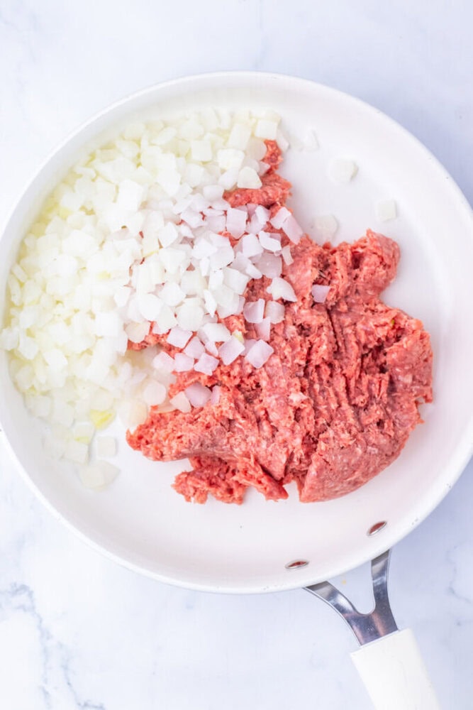 Raw ground beef and chopped onions in a white skillet, ready to be cooked, on a light marble surface.