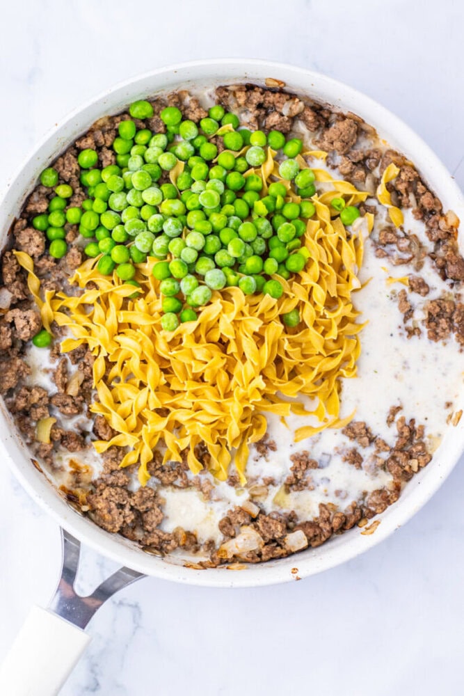 A skillet filled with cooked ground beef, green peas, egg noodles, and a creamy white sauce on a marble countertop.