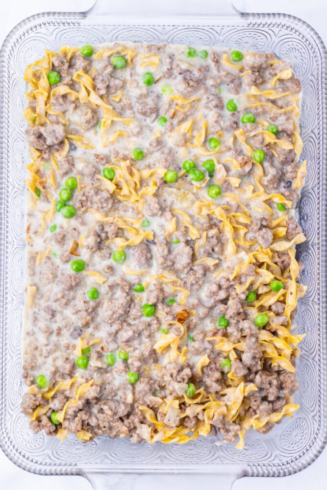 A glass baking dish filled with a casserole made of egg noodles, ground meat, green peas, and a creamy sauce.