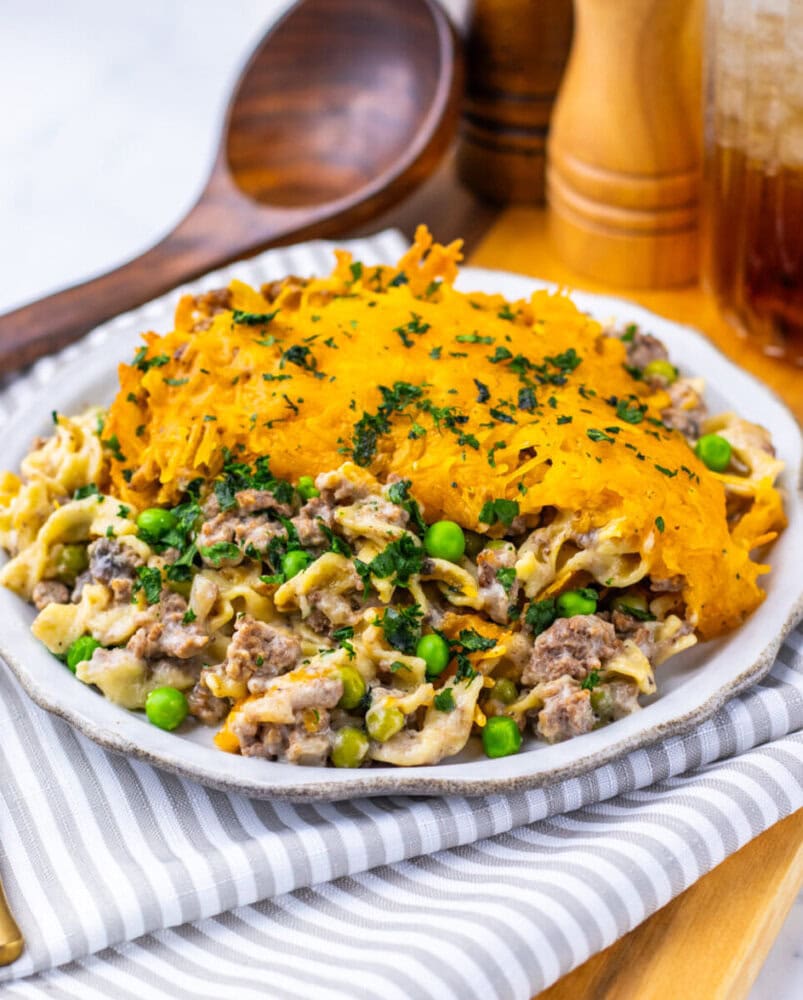 A plate of ground beef and noodle casserole with peas, topped with melted cheddar cheese and parsley, placed on a striped cloth next to pepper mills and a wooden spoon.