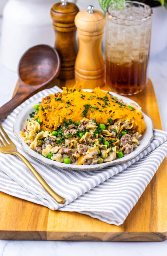 A plate of casserole topped with mashed sweet potatoes, ground meat, peas, and herbs, served with a fork on a striped napkin beside a drink and salt and pepper shakers.