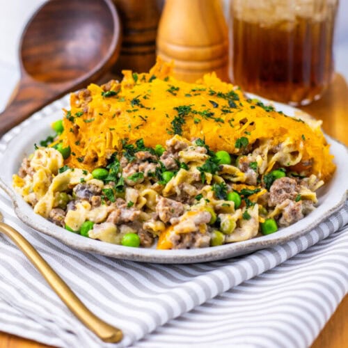A plate of casserole topped with melted cheese, containing ground beef, peas, and noodles, sits on a striped napkin with a fork, wooden spoon, and condiments in the background.