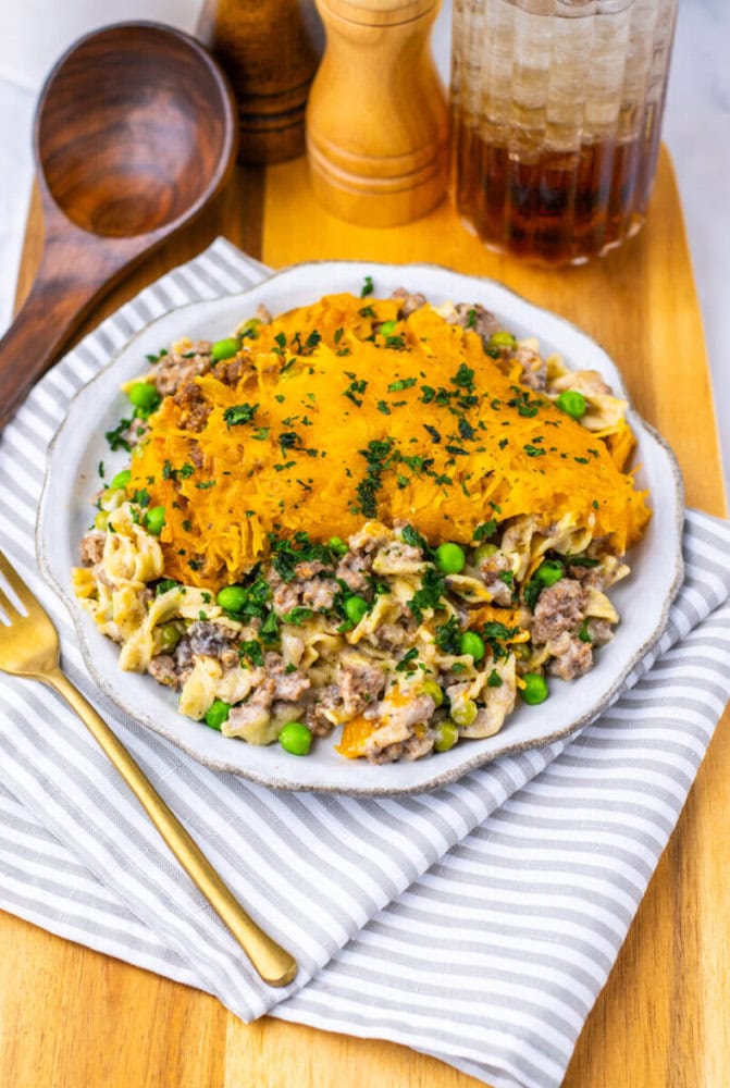 A plate of shepherd&rsquo;s pie with ground meat, peas, egg noodles, and a layer of mashed sweet potatoes, garnished with parsley, sits on a striped napkin beside utensils and condiments.