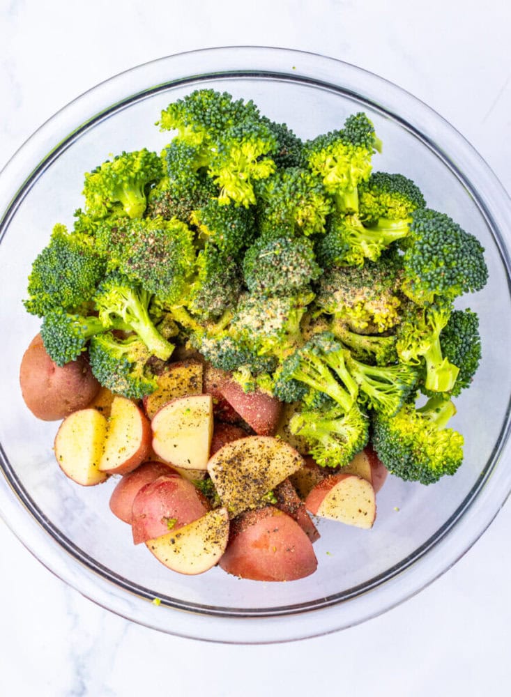 A glass bowl containing chopped red potatoes and broccoli florets, seasoned with black pepper and spices, on a white surface.
