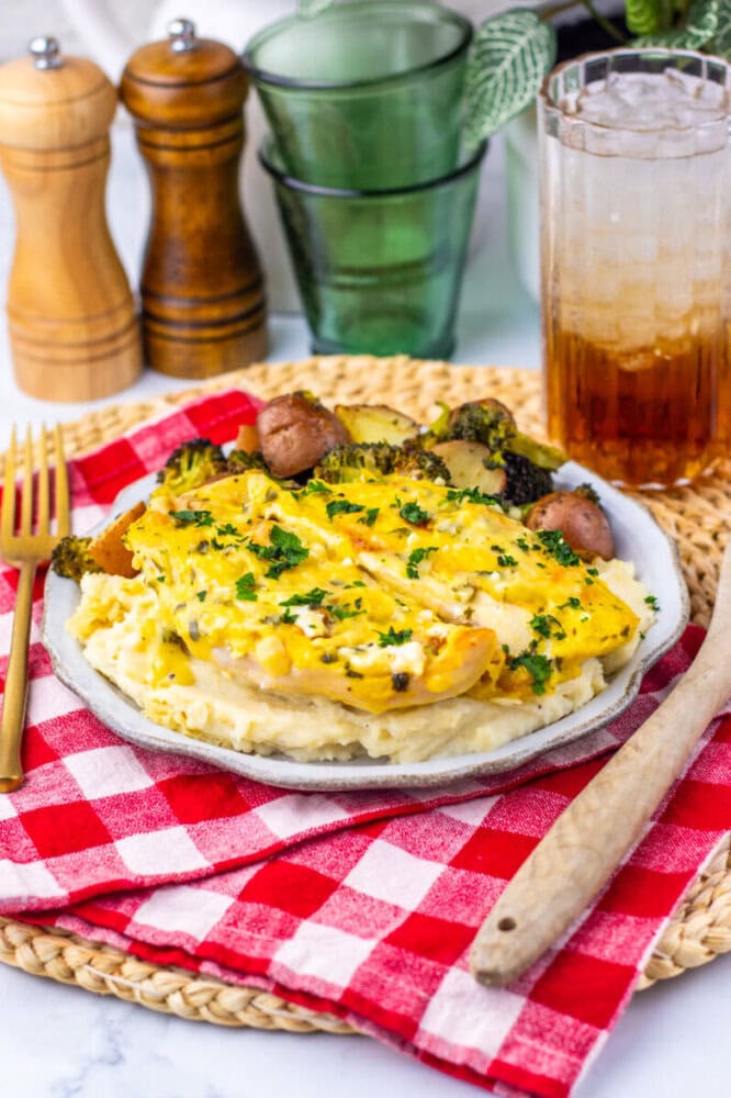 A plate of cheesy chicken and vegetables served over mashed potatoes, placed on a red and white plaid napkin with a fork, knife, and iced drink nearby.