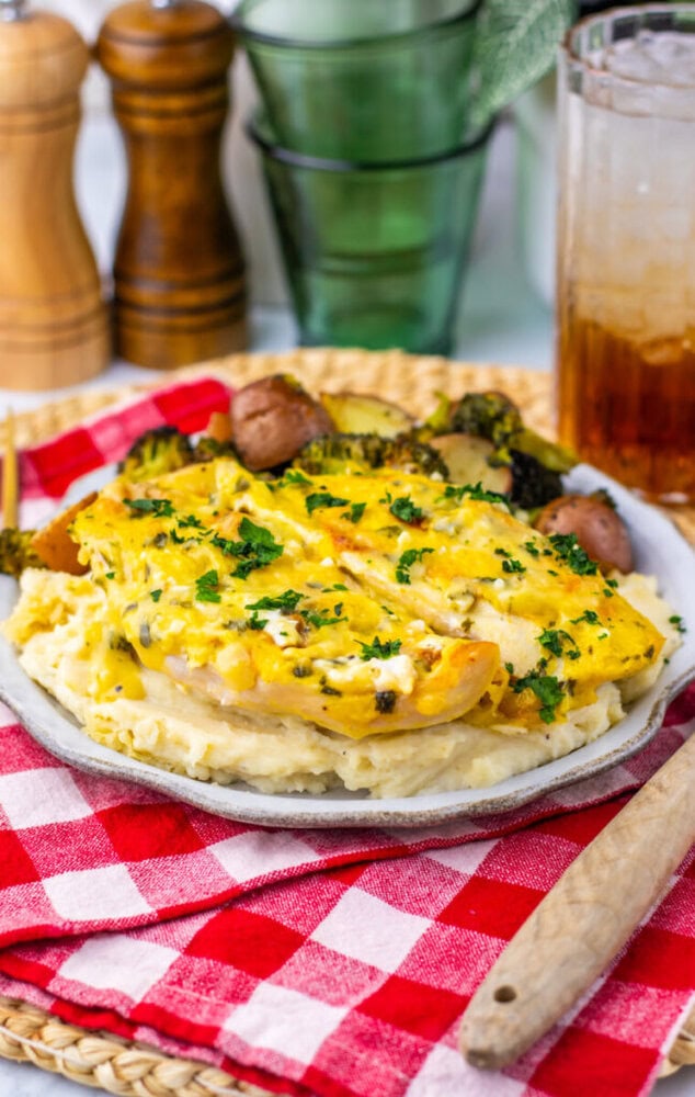 A plate of creamy baked chicken over mashed potatoes with roasted potatoes and broccoli, set on a red checkered napkin beside a glass of iced drink and wooden salt and pepper shakers.