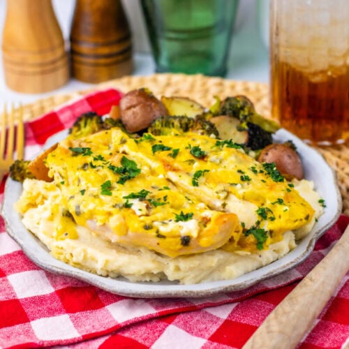 A plate of baked chicken with melted cheese on mashed potatoes, served with roasted vegetables, on a red checkered napkin with a drink and utensils nearby.