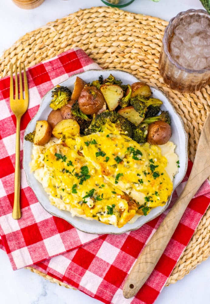 A plate of baked fish topped with cheese and herbs, served with roasted potatoes, broccoli, and a side of mashed potatoes on a red checkered napkin.