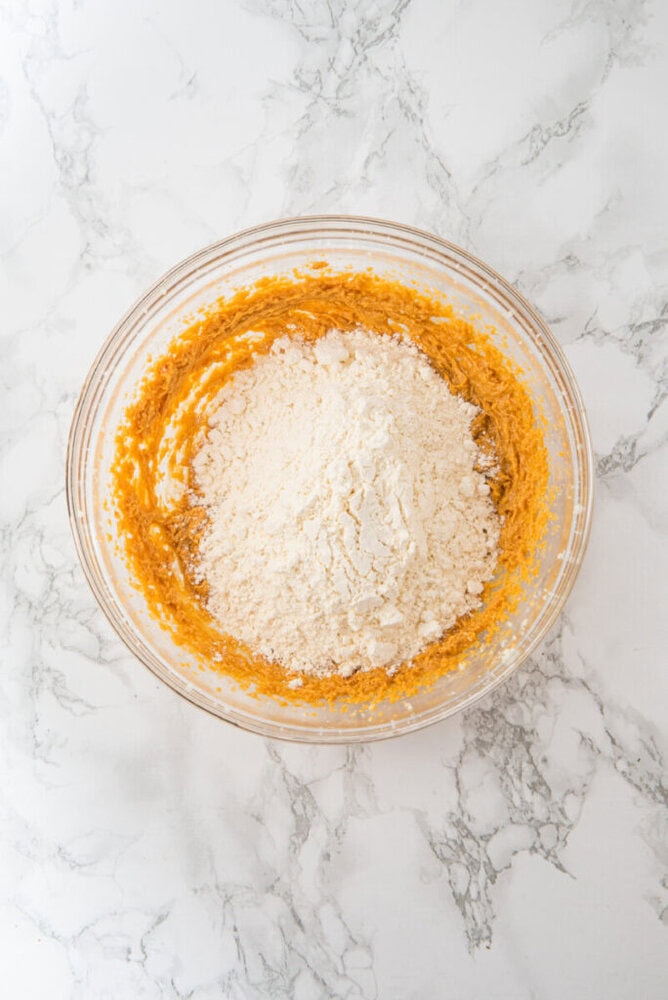A glass mixing bowl containing a partially mixed combination of orange batter and white flour on a marble countertop.