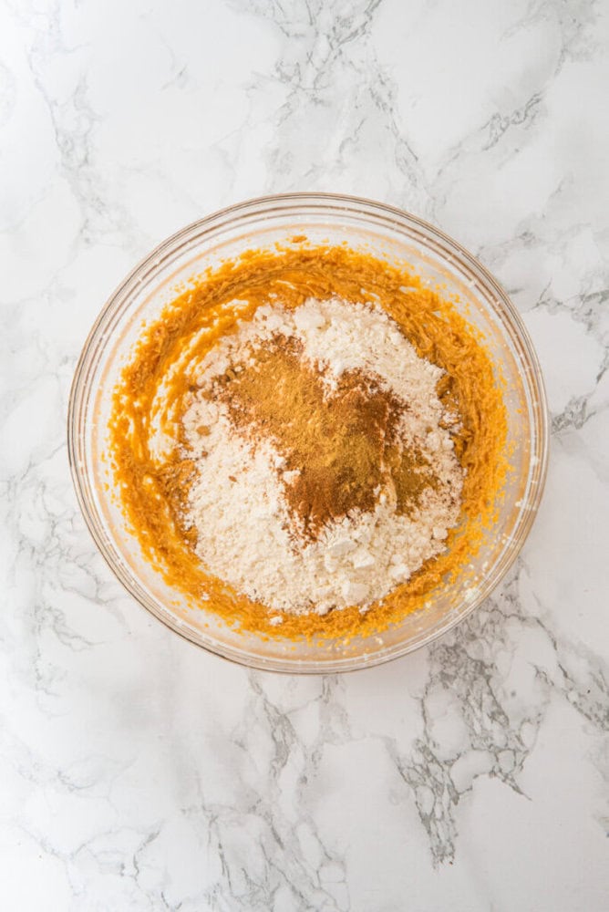 A glass mixing bowl containing a partially mixed batter with flour and spices on a marble surface.