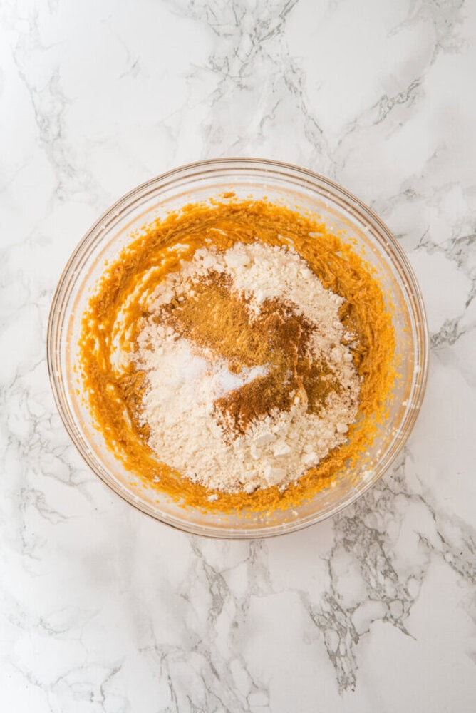 A glass mixing bowl containing a partially mixed blend of flour, spices, and wet ingredients on a white marble surface.
