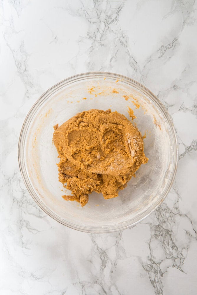 A glass bowl containing partially mixed brown cookie dough sits on a white marble surface.