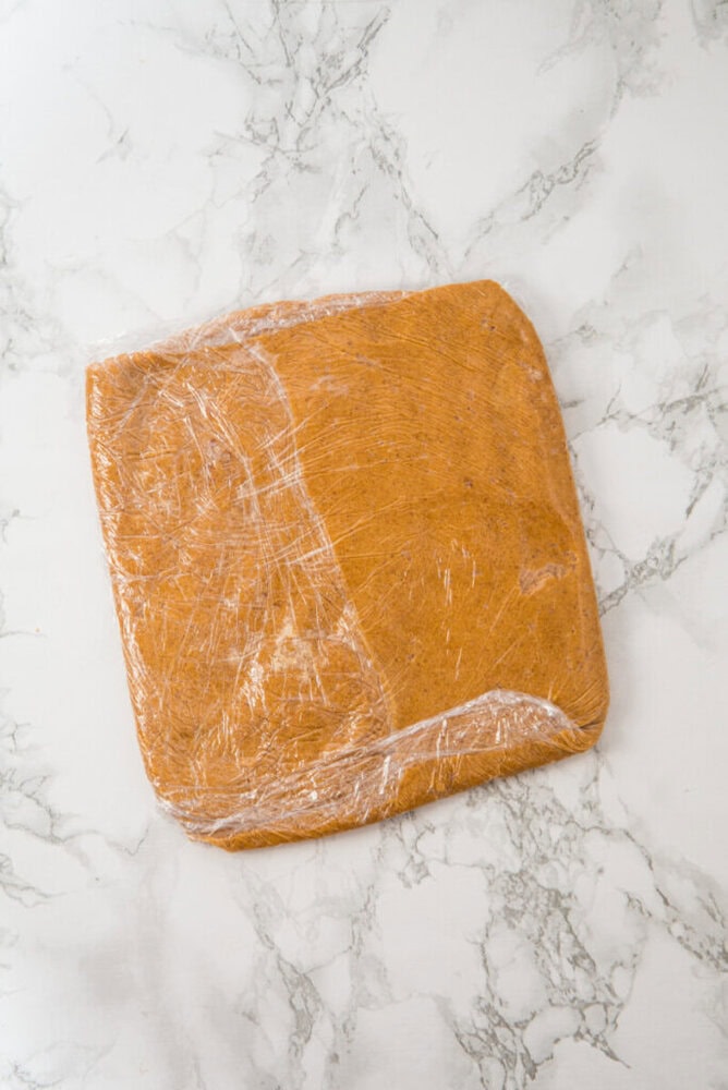 A square slab of brown dough wrapped in plastic wrap sits on a white marble surface.