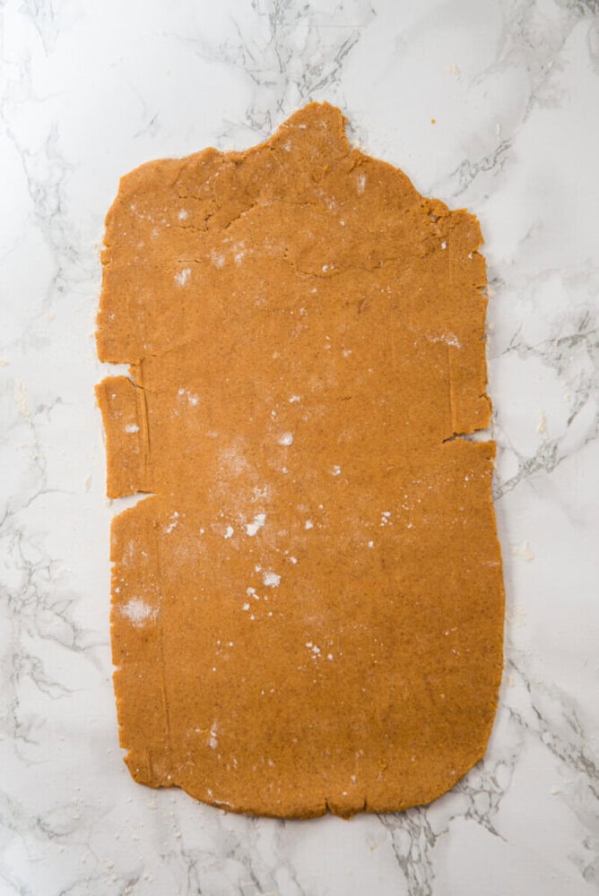 A sheet of rolled-out brown dough with uneven edges on a white marble surface, lightly dusted with flour.