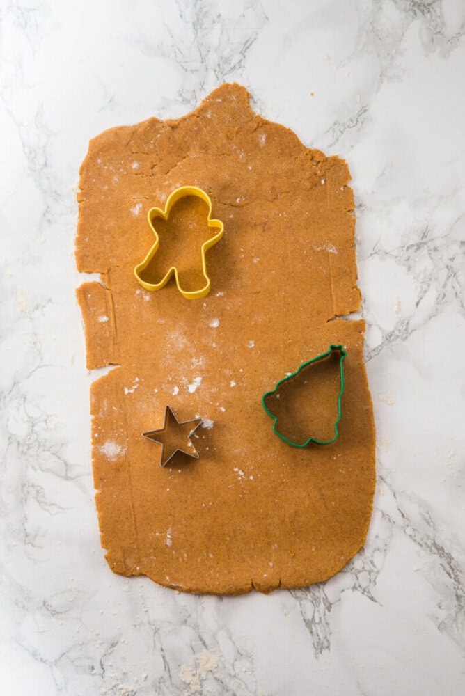Rolled-out gingerbread dough on a marble surface with star, Christmas tree, and gingerbread person cookie cutters placed on top.