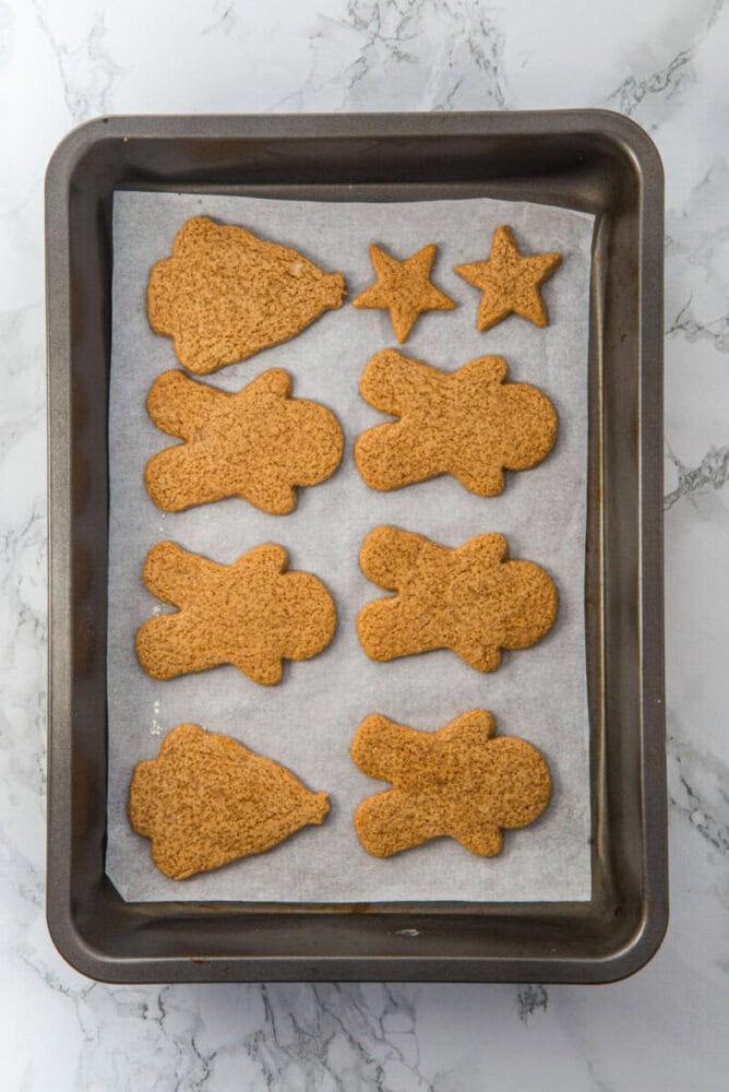 A baking tray lined with parchment paper holds eight gingerbread cookies in tree, star, and gingerbread person shapes on a marble countertop.