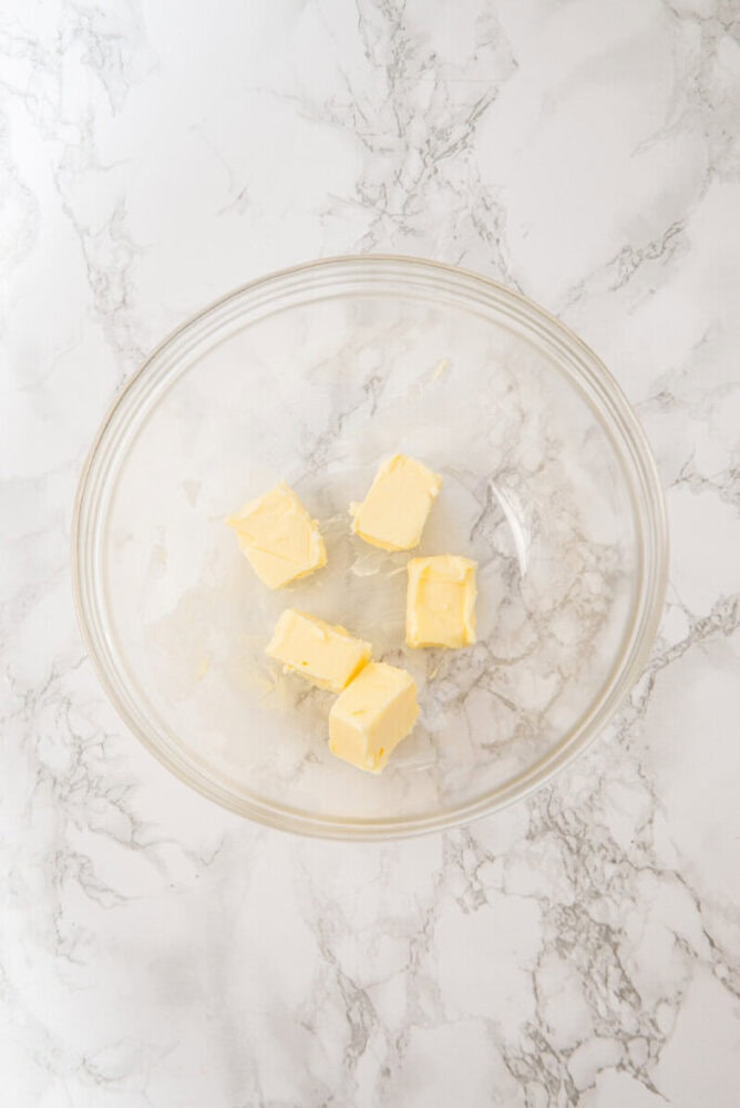 A glass bowl containing five cubes of butter sits on a white marble surface.