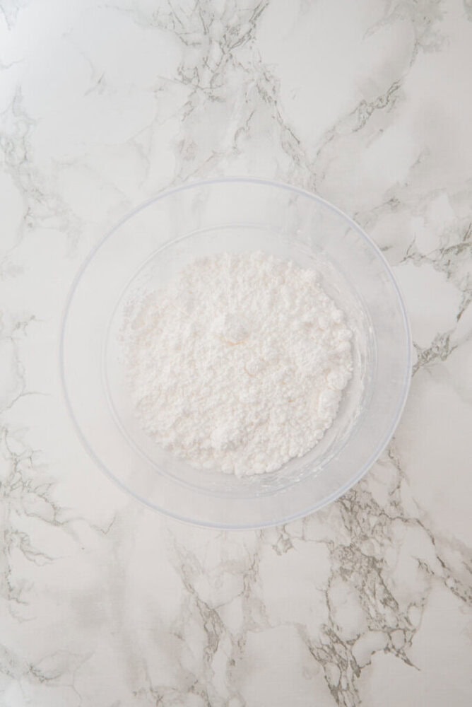 Clear mixing bowl filled with white flour and small clumps, placed on a white marble countertop.