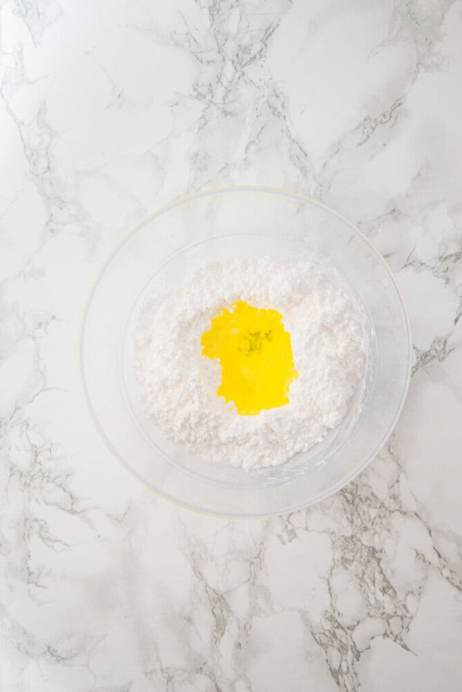 A clear bowl with flour and a pool of liquid egg in the center sits on a white marble surface.