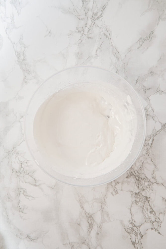 A clear bowl filled with thick, white whipped cream sits on a white marble surface.