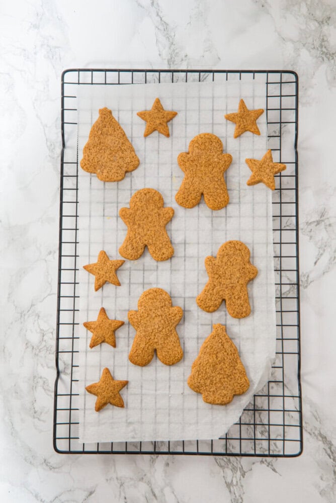 A cooling rack holds gingerbread cookies in the shapes of stars, trees, and gingerbread people on a sheet of parchment paper over a marble surface.