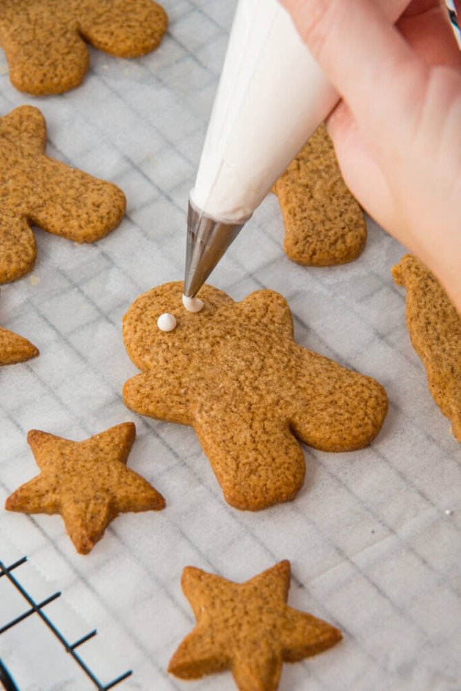 A hand pipes white icing onto a gingerbread person cookie on a cooling rack, surrounded by other gingerbread cookies in star and person shapes.