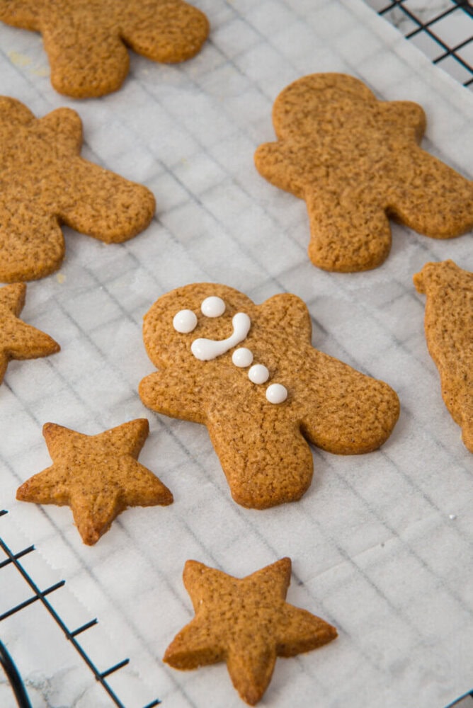 Gingerbread cookies in star and person shapes, with one decorated with white icing, are cooling on a wire rack lined with parchment paper.
