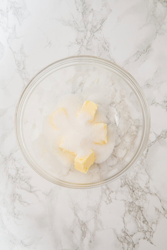 A glass bowl containing several pieces of butter and a mound of granulated sugar on a white marble surface.